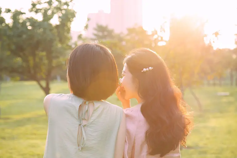 Two women enjoy a sunny day outdoors, whispering and smiling.