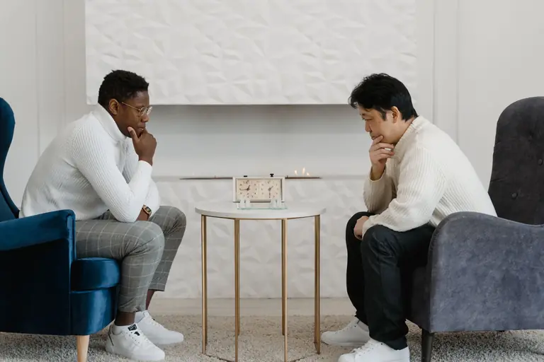 Two men engage in a thoughtful chess game indoors with a chess clock on a round table.