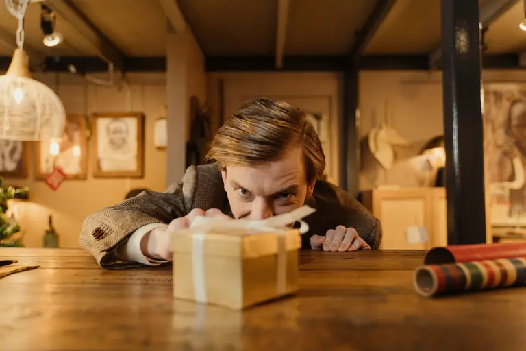 A man eagerly looks at a wrapped present on a table in a cozy room filled with warm lighting and decorations.