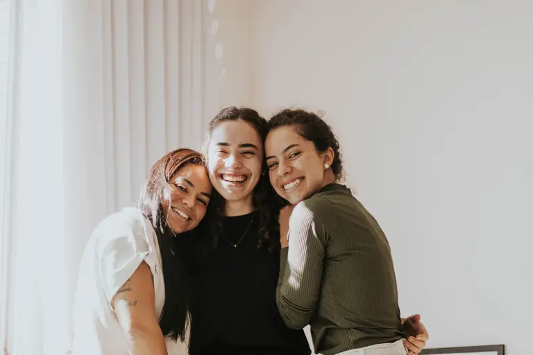 Three smiling friends hugging indoors, creating a warm and joyful atmosphere.