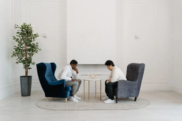 Two men seated in armchairs focus on a strategy board game in a modern, minimalist room.