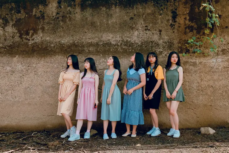 Six young women in summer dresses stand against a rustic wall in an outdoor setting.